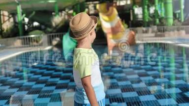 时髦的旅行者，戴着太阳镜和帽子的<strong>婴儿</strong>，指着<strong>游泳池</strong>附近的操场滑梯